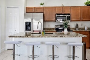 Una cocina con encimera de granito, electrodomésticos de acero inoxidable, armarios de madera y cuatro taburetes blancos.