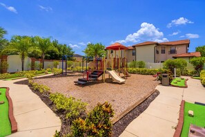 A playground with a slide and climbing structures, surrounded by a landscaped garden with palm trees and shrubs. - 4BR Villa|Pool•Game Room•Free BBQ|Disney (Clermont)
