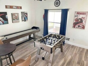 A room with a foosball table, a bench, and a table with chairs. There are posters on the wall and a window with curtains.