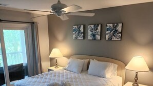 Un dormitorio con cama, dos mesitas de noche con lámparas, ventilador de techo y tres obras de arte colgadas en la pared.