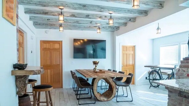 Un comedor con una mesa de madera, sillas y un gran cuenco de madera como adorno central. También hay taburetes de madera, un televisor montado en la pared y una bicicleta al fondo.