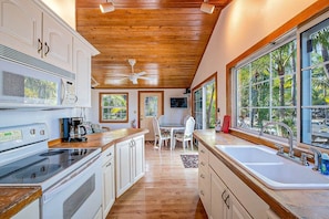 A modern kitchen with wooden ceiling beams, a dining area with a table and chairs, and large windows offering a view of palm trees.