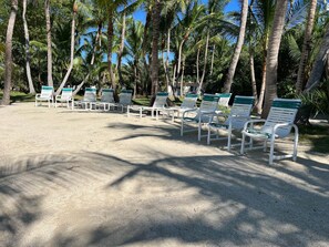 A beachside seating area with white and green lounge chairs arranged on a sandy surface, surrounded by tall palm trees.