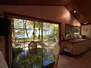 Una habitación con una mesa y sillas de vidrio, una zona de cocina con lavaplatos y armarios, y vistas al exterior a través de grandes ventanales.