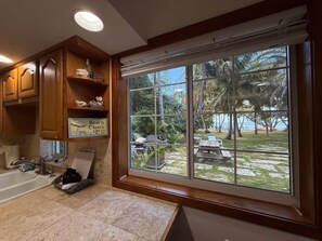 Un baño con armarios de madera, un lavamanos y un ventanal con vista a una playa y palmeras.