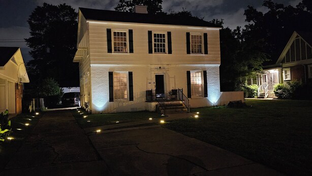 A two-story white house with a front porch, illuminated windows, and a small front yard with decorative lights.