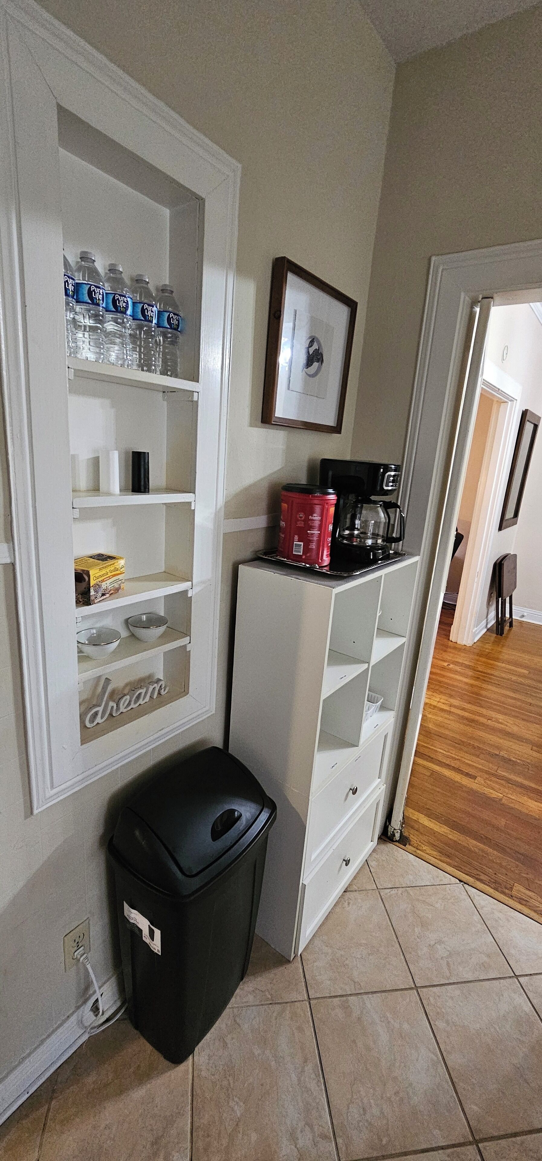 A kitchen with a white cabinet containing water bottles, a coffee maker, and a trash bin.