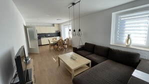 A modern living room with a sofa, a coffee table, and a TV. There is a dining area with a table and chairs. The room has a large window with blinds. - Vacation apartment in the heart of Saxony (Leisnig)