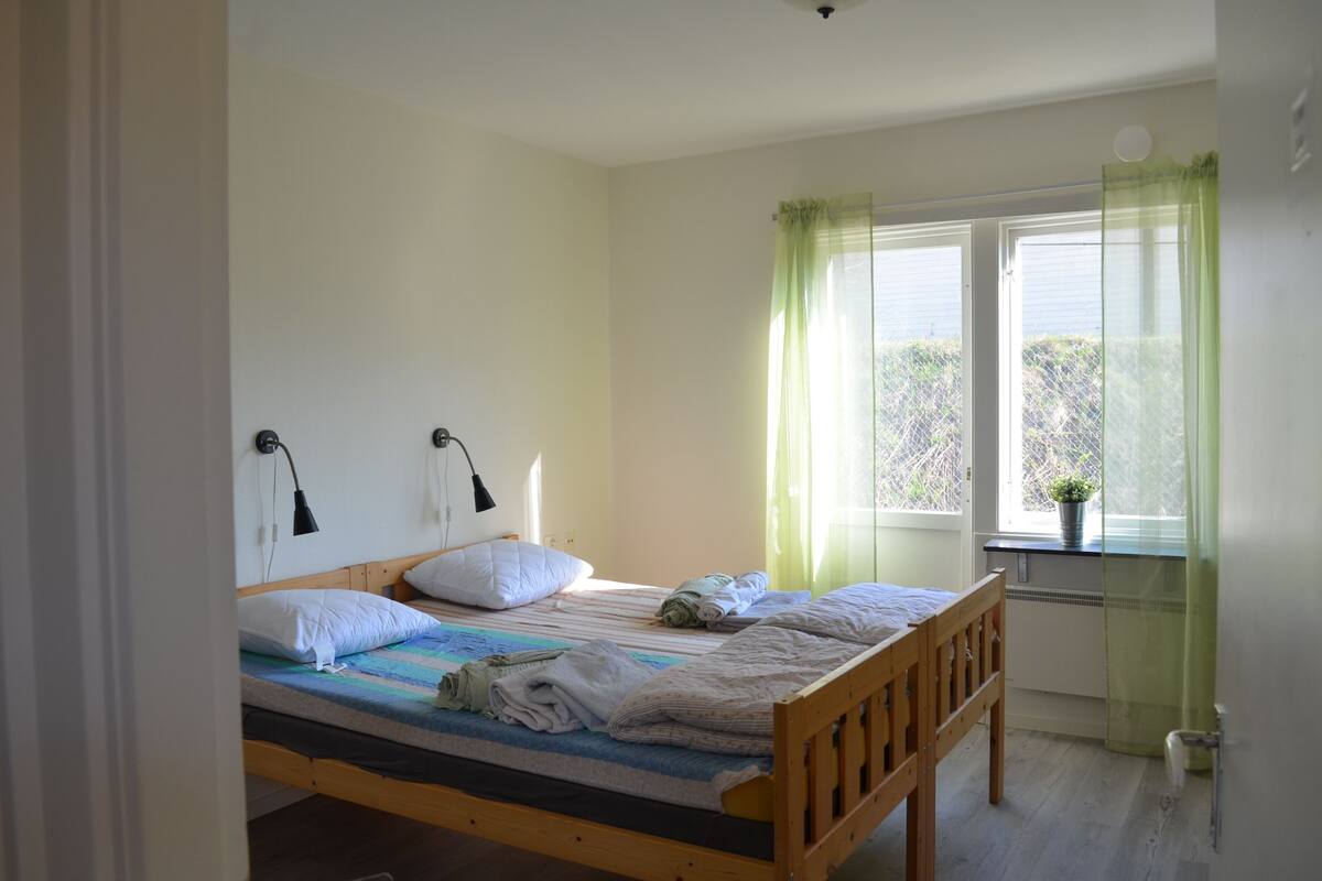Ein Einzelbett mit Holzrahmen, zwei Nachttischlampen, ein Fenster mit durchsichtigen Vorhängen und ein kleiner Topfpflanzen auf der Fensterbank.