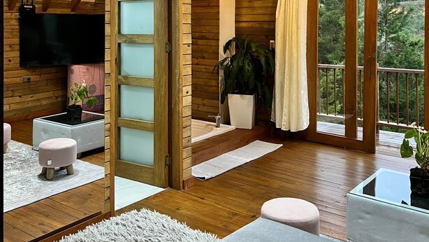 A spacious room with wooden beams, a large window, a flat-screen TV, a sofa, a coffee table, and a potted plant. - Quimera Ecolodge Cabana - 7 min JMC Airport (Antioquia)