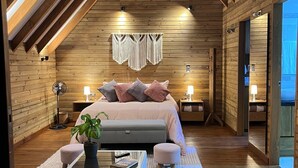 A cozy attic bedroom with a sloped ceiling, wooden walls, and a large bed with pillows. There are two bedside tables with lamps, a floor lamp, and a small table with a plant and books. - Quimera Ecolodge Cabana - 7 min JMC Airport (Antioquia)