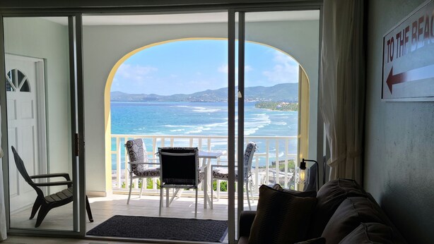 A room with a sliding glass door leading to a balcony with a view of the sea and mountains. There is a couch, a table, and a chair inside the room.