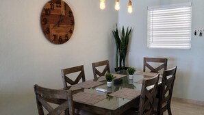 A dining area with a wooden table, six chairs, a round wooden clock, a hanging light fixture, and a window with blinds.