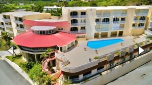 A multi-story hotel with a rooftop pool, a curved red-roofed structure, and a landscaped area with plants.