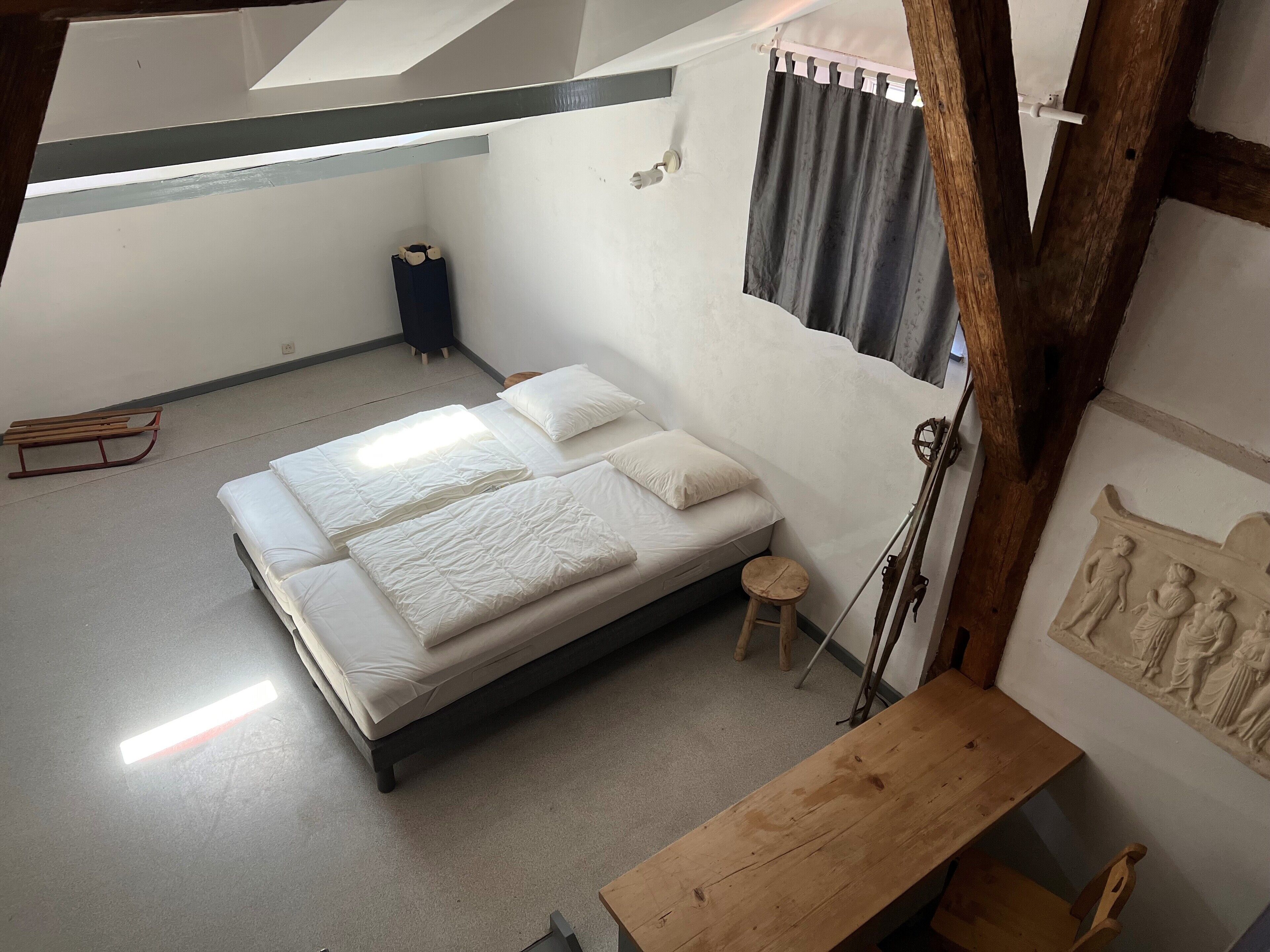 Un dormitorio con una cama, un taburete pequeño, un banco, una guitarra y una pared con un relieve escultórico.