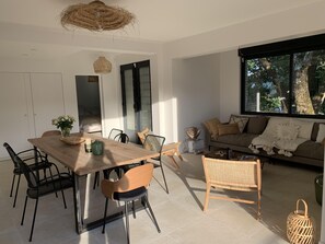 A modern living room with a wooden dining table, black chairs, a sofa with cushions, and a large window offering a view of trees.