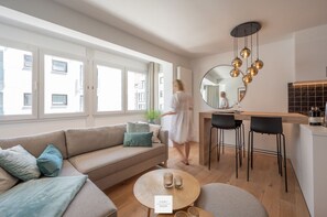 En moderne stue med sofa, rundt sofabord og et køkkenområde med barstole.