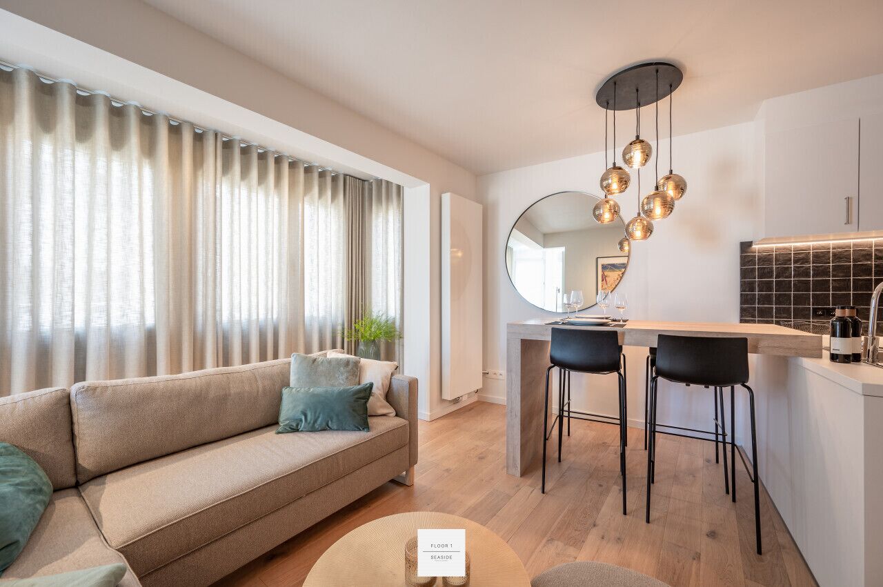 A modern living room with a sofa, a dining area with two chairs, a kitchen with a bar, and a large mirror.