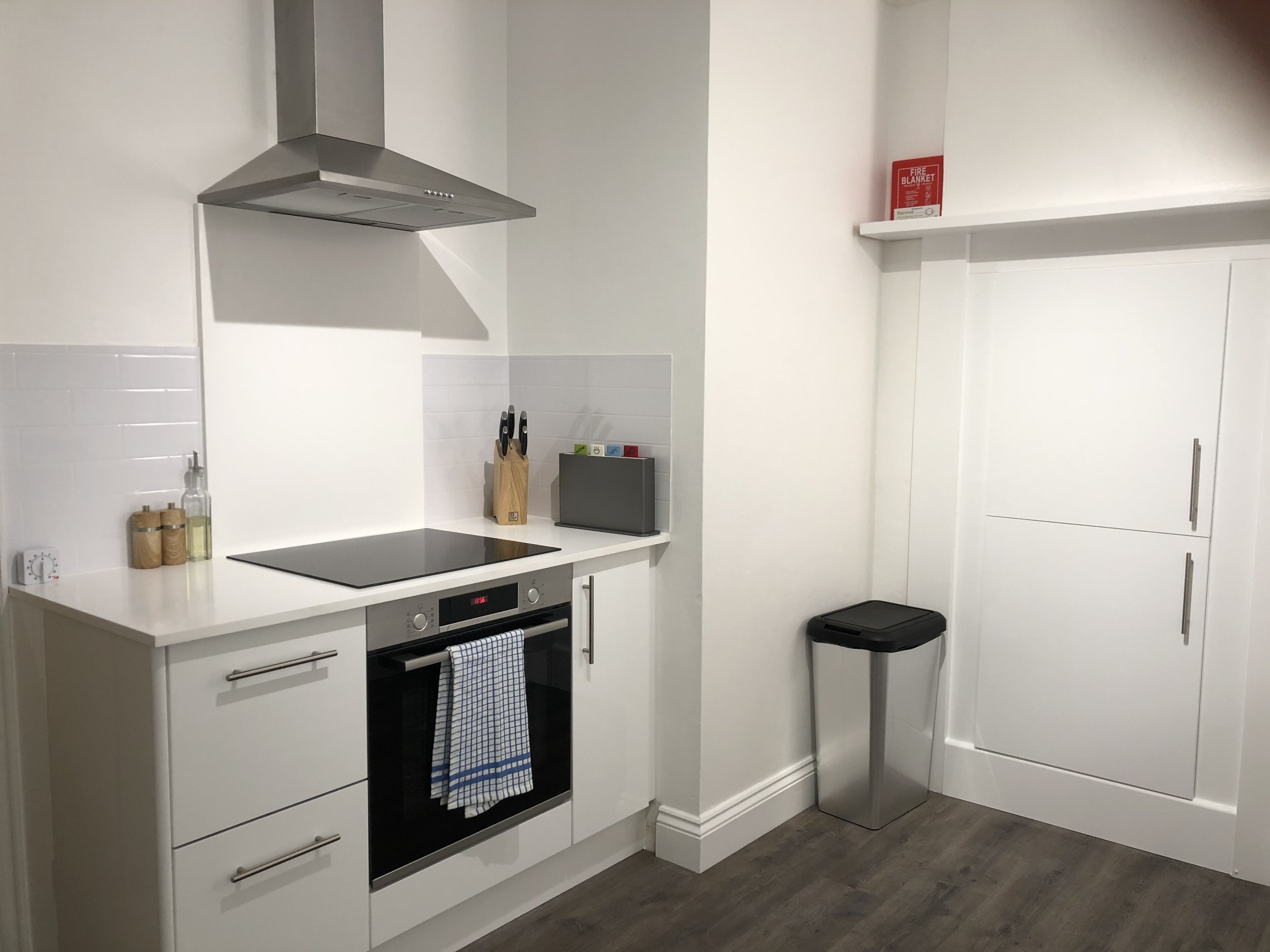 A modern kitchen with white cabinetry, a stainless steel range hood, a built-in oven, and a black trash bin.