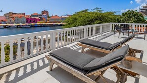 Een balkon met twee houten loungestoelen en een tafel, met uitzicht op een haven met kleurrijke gebouwen en boten.