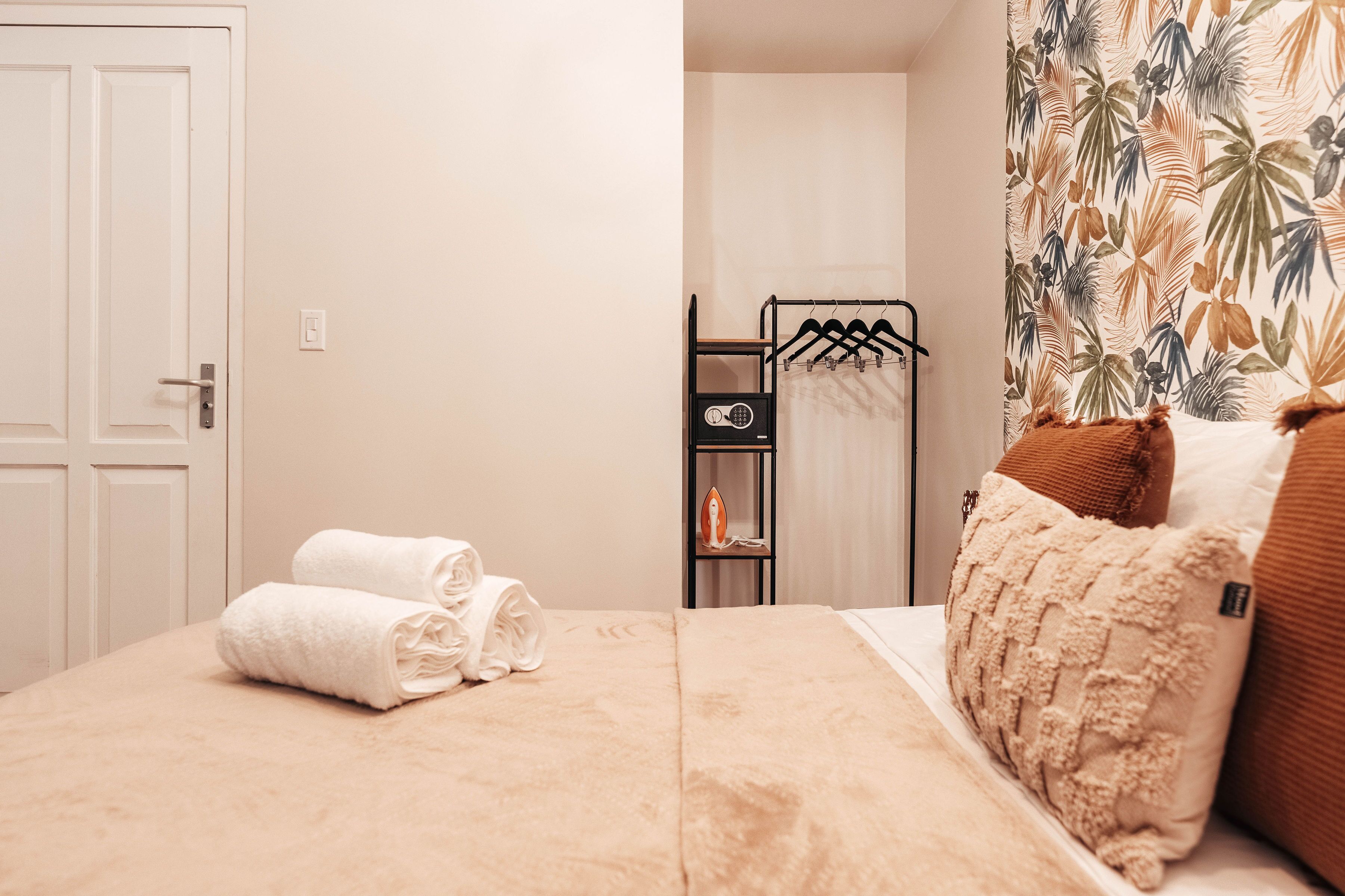 A neatly made bed with rolled towels, a black metal hanger rack, and a small shelf with an iron.