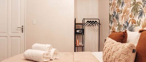 A neatly made bed with rolled towels, a black metal hanger rack, and a small shelf with an iron.