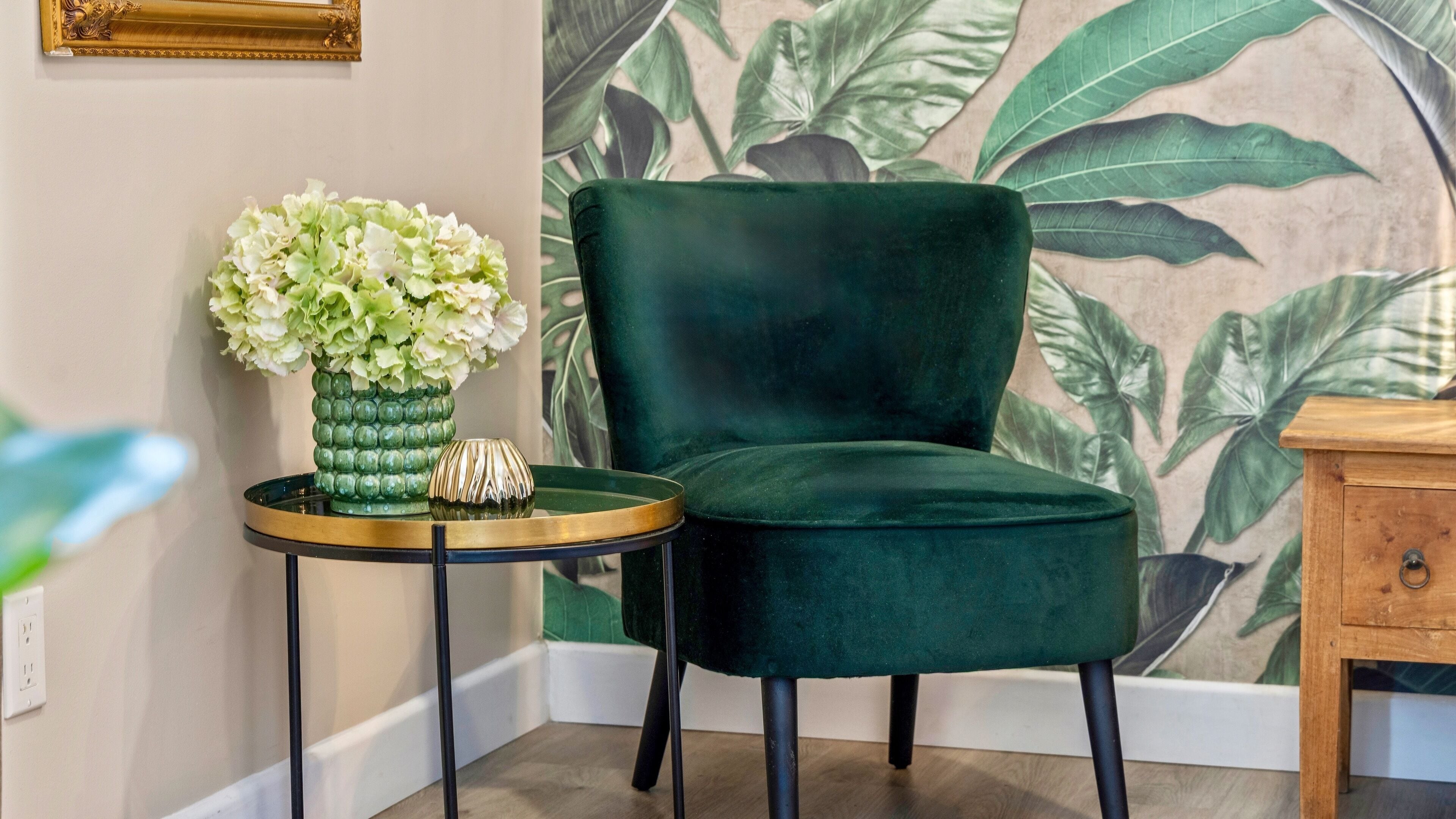 A corner of a room with a green velvet chair, a small round table with a green vase and a decorative bowl, and a wall with a tropical leaf pattern.