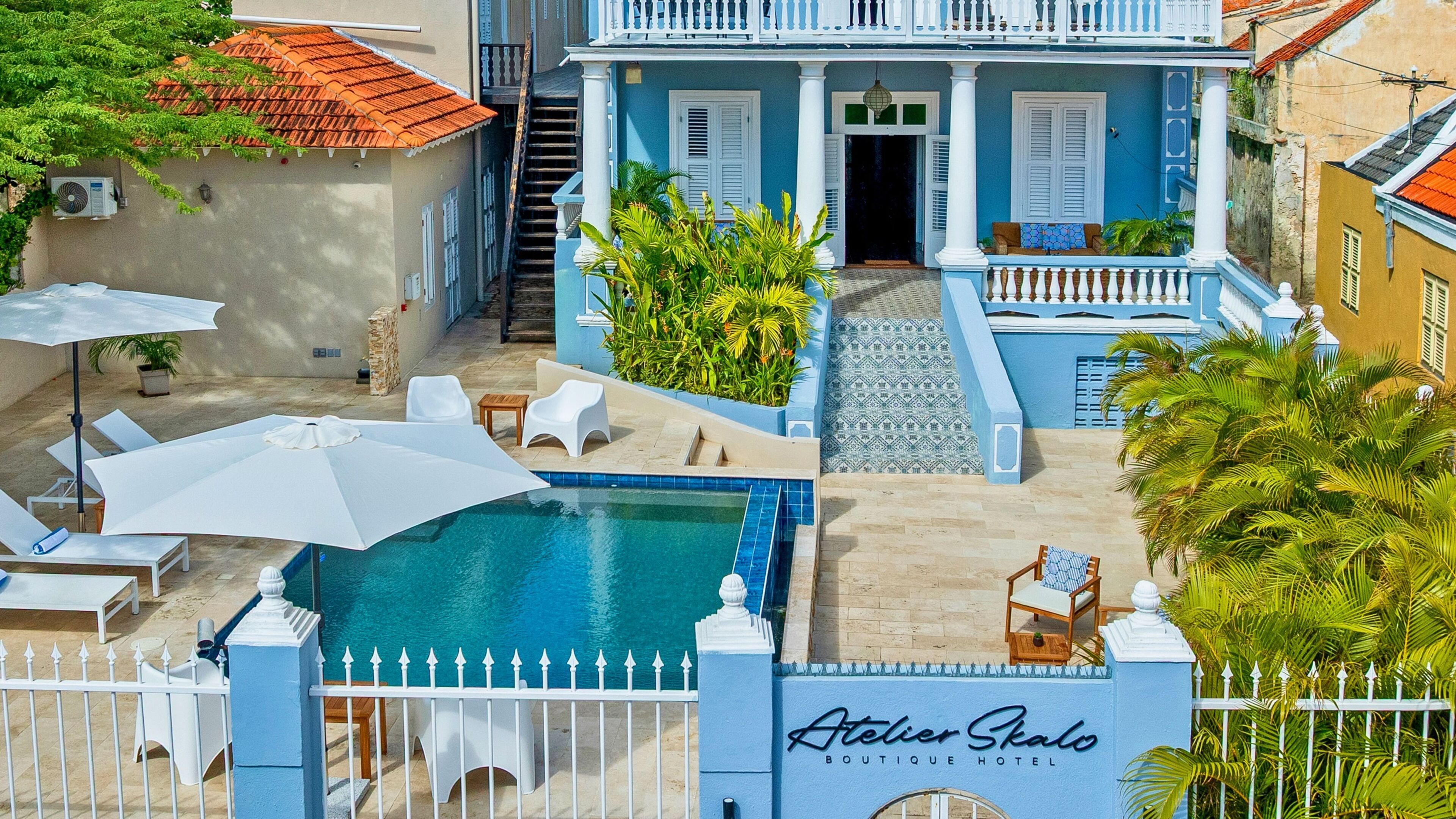 Zona de piscina con sillones de playa y sombrillas, una escalera que lleva a un edificio de fachada azul, y un letrero que dice 'Atelier Skalo Boutique Hotel'.