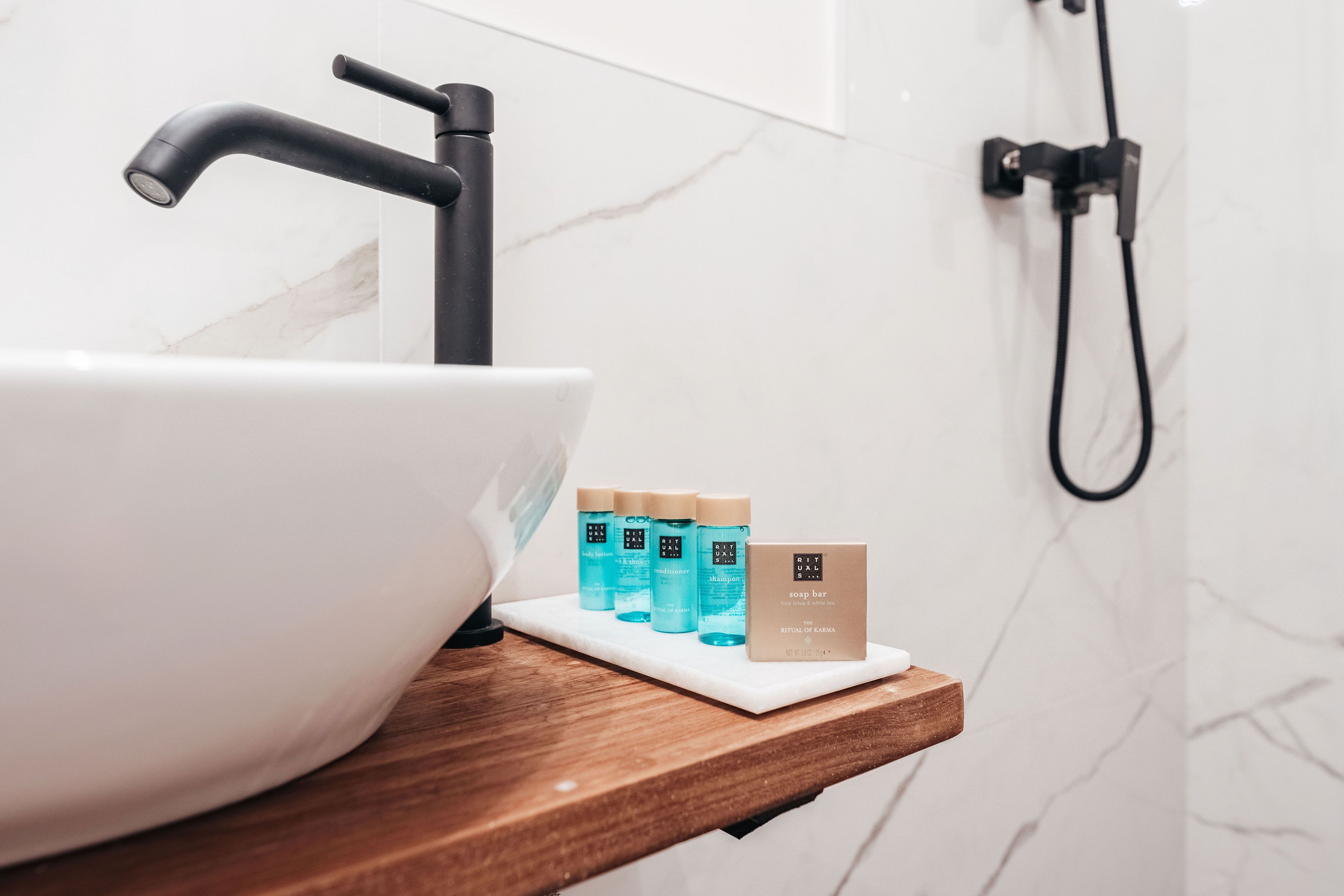Un baño con un lavabo de mármol blanco, un grifo negro y un estante de madera con tres botellas de champú y una barra de jabón.