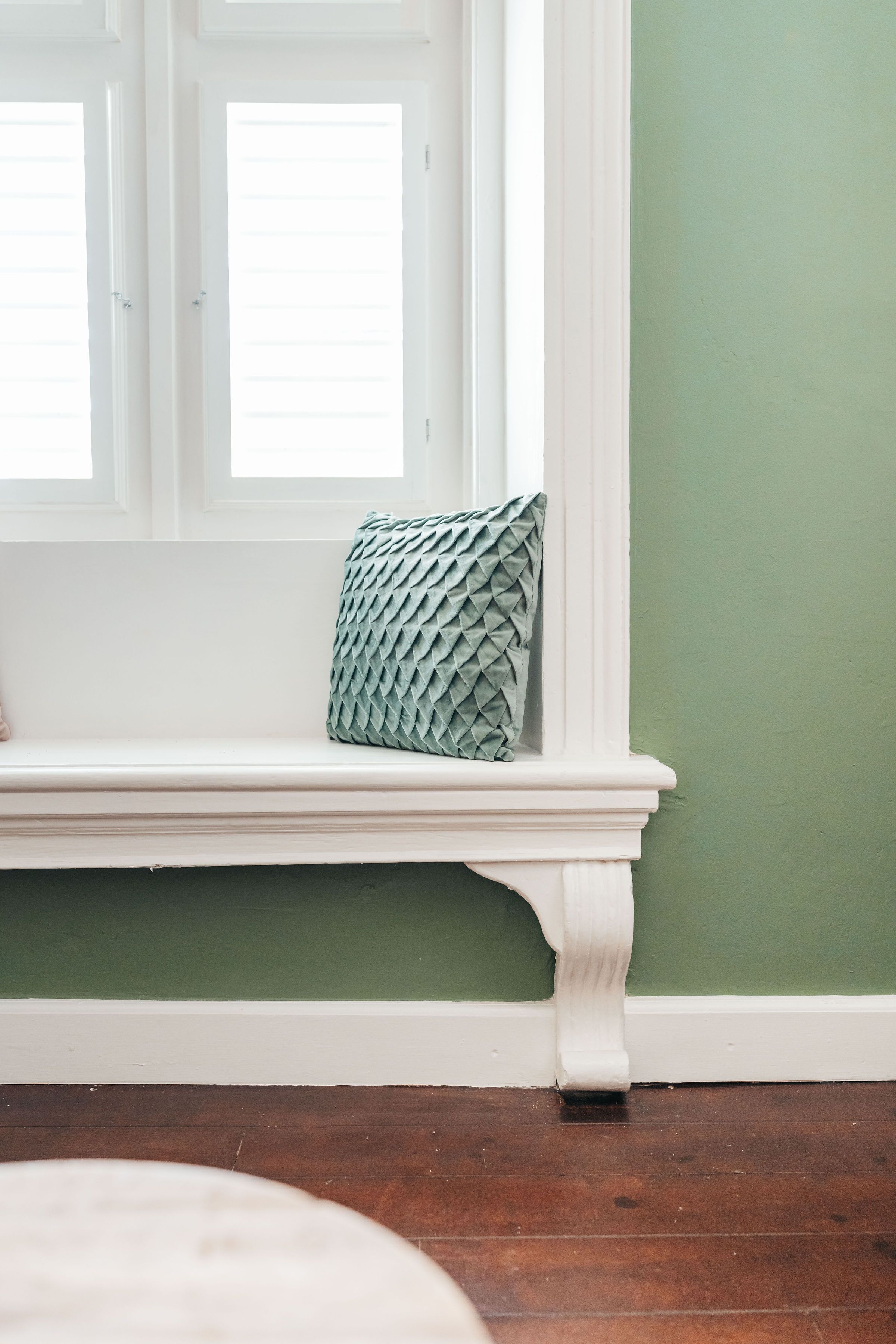 Une pièce avec un banc blanc adossé à un mur vert, une fenêtre avec des stores et un plancher en bois.