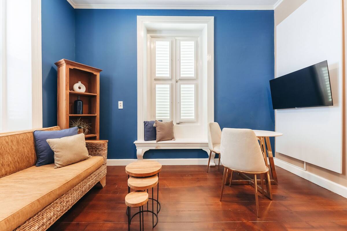 Un salon avec un mur bleu, un banc blanc, une bibliothèque en bois, un canapé, une table ronde en bois et un téléviseur fixé au mur.