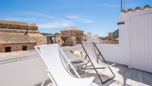 Une terrasse sur le toit, avec des chaises blanches, une petite table ronde ornée d’un vase de fleurs, et une vue sur des bâtiments historiques et une tour en arrière-plan.