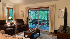 Een woonkamer met een bank, fauteuils, een salontafel en een televisie. Er is een groot raam met gordijnen, een balkon met tuinmeubilair, en een vloerkleed.