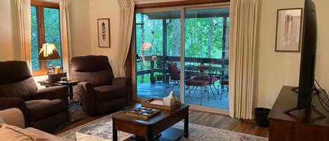 A living room with a sofa, armchairs, a coffee table, and a television. There is a large window with curtains, a balcony with outdoor furniture, and a rug on the floor.