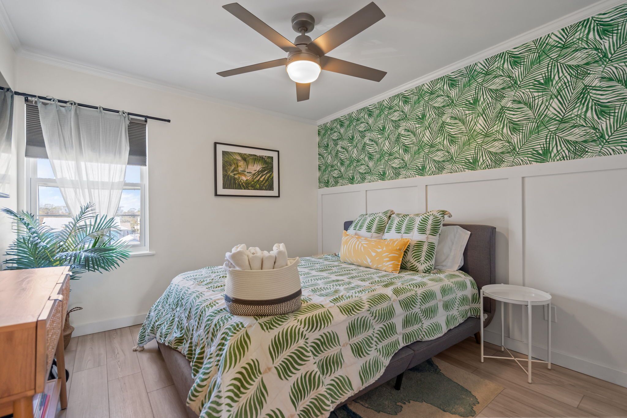 Une chambre avec un lit, un ventilateur de plafond, une fenêtre avec des rideaux légers, un mur orné d’un motif de feuilles, une petite table et une plante.