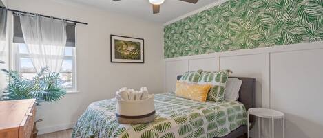 Une chambre avec un lit, un ventilateur de plafond, une fenêtre avec des rideaux légers, un mur orné d’un motif de feuilles, une petite table et une plante.