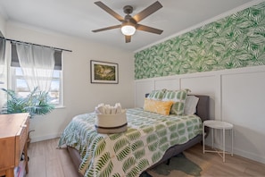A bedroom with a bed, a ceiling fan, a window with sheer curtains, a wall with a leaf pattern, a small table, and a plant.