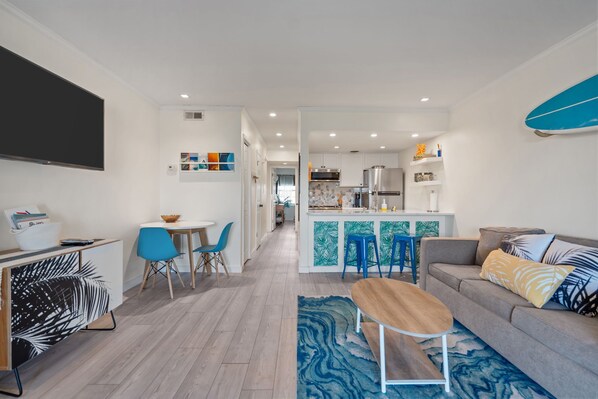 A modern living room with a sofa, coffee table, and a TV. There is a kitchen area with a fridge, microwave, and a dining table with chairs.