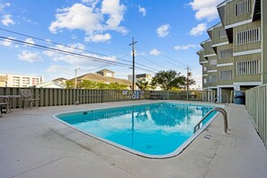 A swimming pool with a metal safety railing, surrounded by a concrete deck, with buildings and a clear sky in the background.