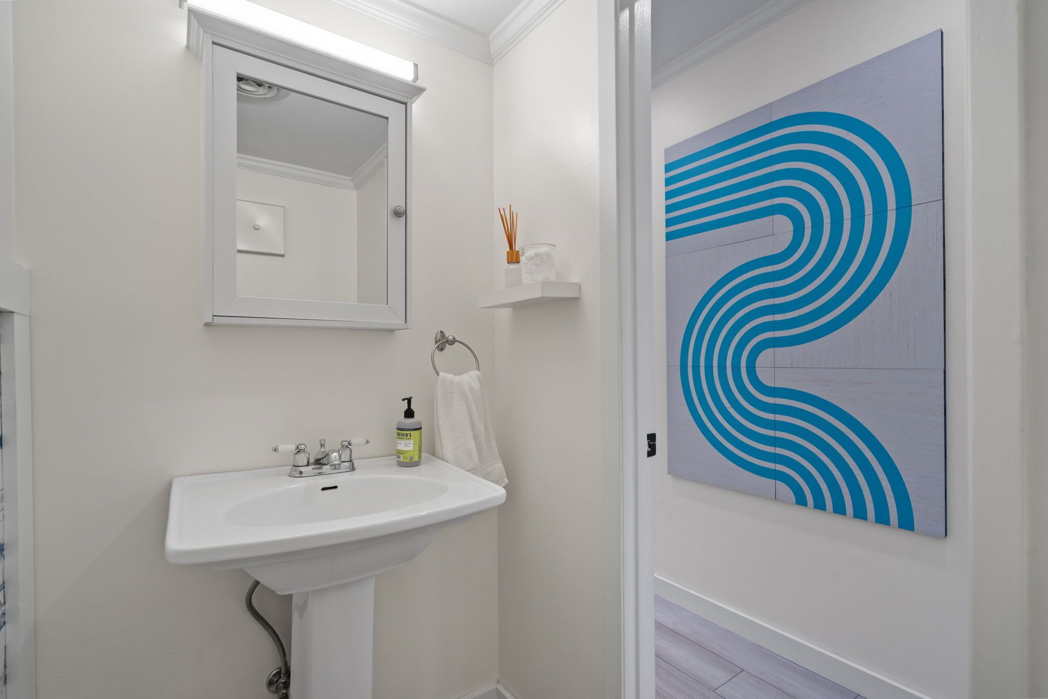 Un bagno con un lavandino bianco, uno specchio e un quadro astratto blu appeso al muro.
