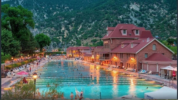 A swimming pool area with multiple pools, a building with a red roof, and a mountainous backdrop.