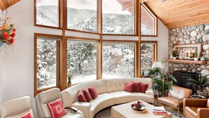 A cozy living room with a curved sofa, a coffee table, and a fireplace. Large windows offer a view of a snowy mountain landscape.