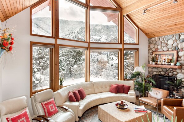 A cozy living room with a curved sofa, a coffee table, and a fireplace. Large windows offer a view of a snowy mountain landscape.