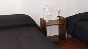 Une petite pièce simple, avec deux lits simples, une table de chevet en bois avec une lampe et une prise électrique au mur.
