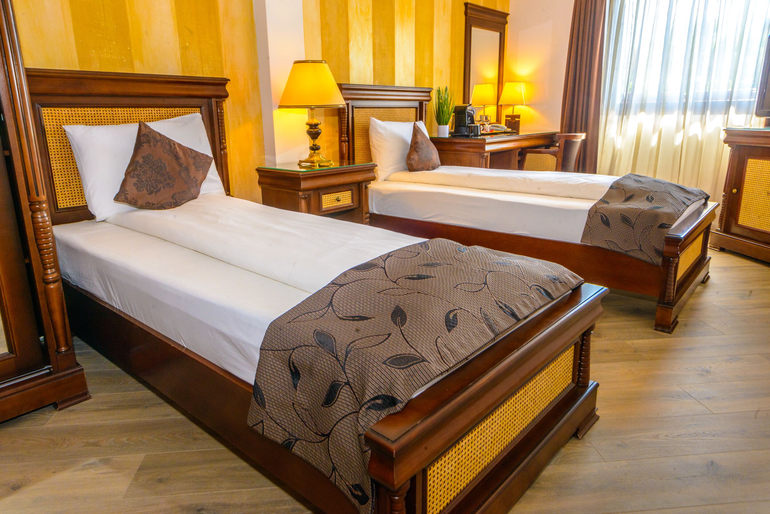 A hotel room with two wooden beds, each with a headboard, bedside tables, lamps, and a small potted plant.