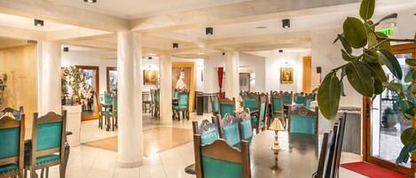 A spacious dining area with high ceilings, wooden chairs with teal upholstery, and a central table with a lamp.