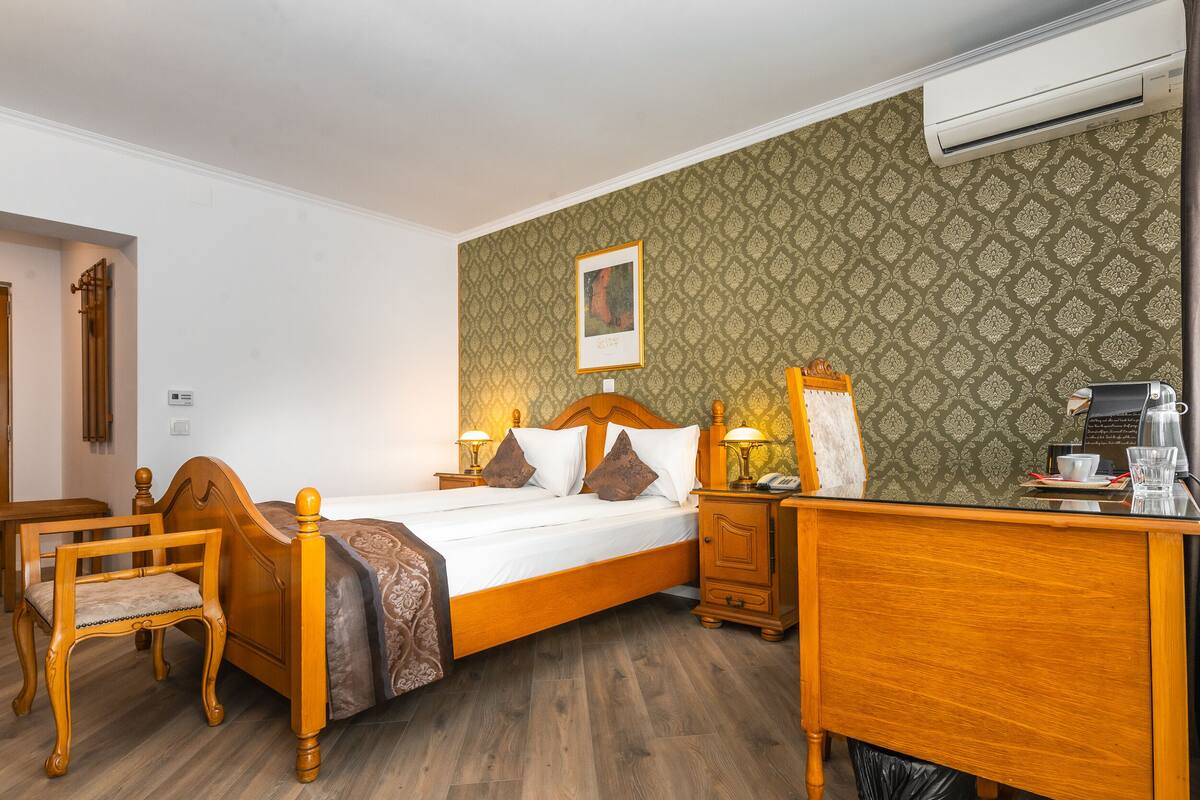 A hotel room with a wooden bed, a desk with a telephone, a chair, and a small table with a cup and a glass.