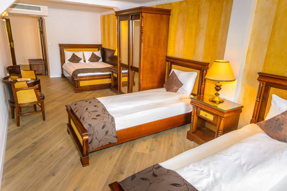 A hotel room with two beds, a sitting area with a chair, a bedside table with a lamp, and a wardrobe.