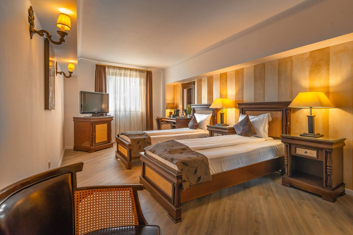 A hotel room with two beds, a television, wooden furniture, and wall-mounted lamps.