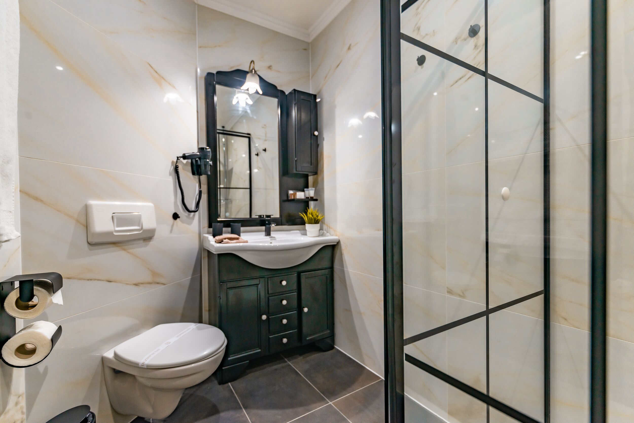 A bathroom with a white toilet, dark green cabinets, a white sink with a mirror above it, and a glass shower enclosure.